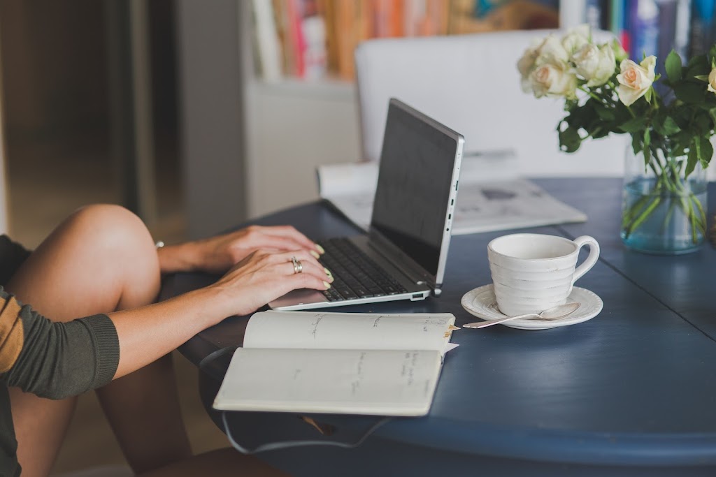 A women writing on laptop with a cup of tea and a book beside.